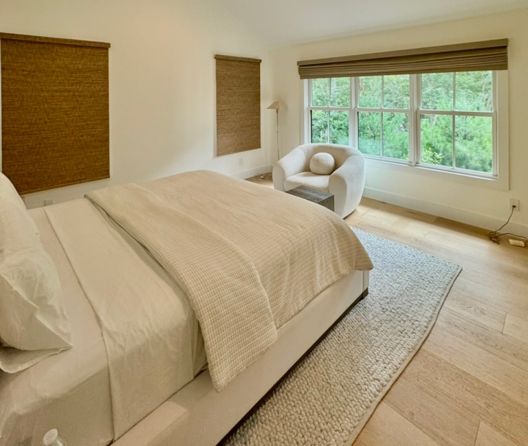 A bright, minimalist bedroom features a neatly made bed with a beige woven blanket and white pillows. An inviting chair sits by the large window adorned with affordable blinds from NYC. The room boasts light wooden floors and a soothing neutral color palette.