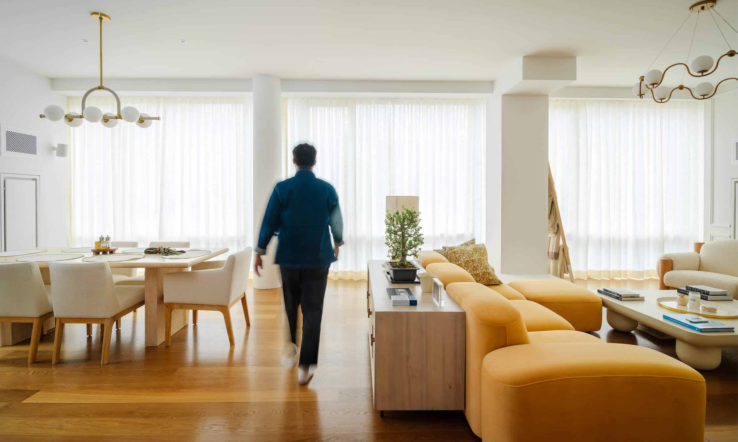 A person walks through a modern, elegantly furnished living room with large windows adorned with custom window treatments NYC. The space features sheer curtains, wooden flooring, a yellow sofa, a dining area with a table and chairs, and unique overhead light fixtures.