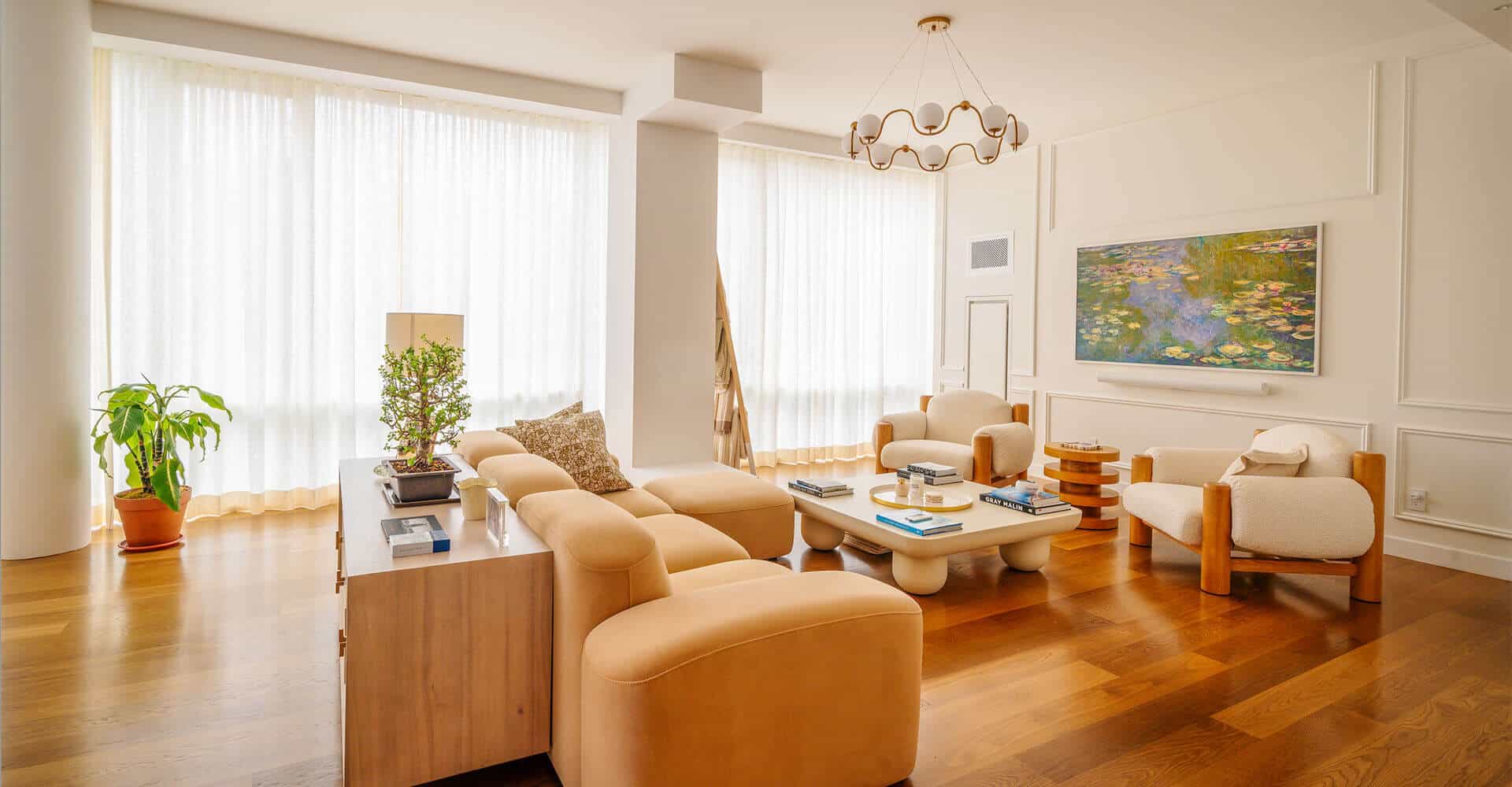 A modern living room with a white sofa, gray cushions, and a black armchair. Floor-to-ceiling custom draperies NYC frame a large window, allowing natural light. The decor includes a lamp, abstract artwork, and a table with pink tulips in a vase.