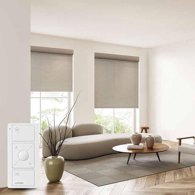 A minimalist interior showcases large windows adorned with beige roller blinds, reflecting the elegance of custom draperies NYC. A wireless remote control rests on a white surface in the foreground.