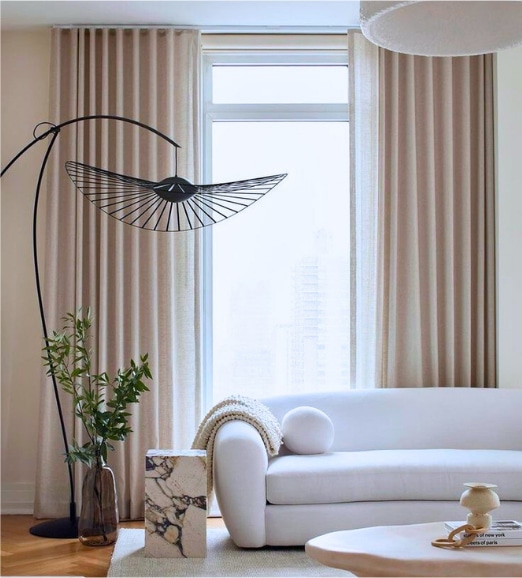 A modern living room with beige curtains, a white sofa, a unique black floor lamp, a small marble side table, a plant in a glass vase, and soft natural light coming through a large window.