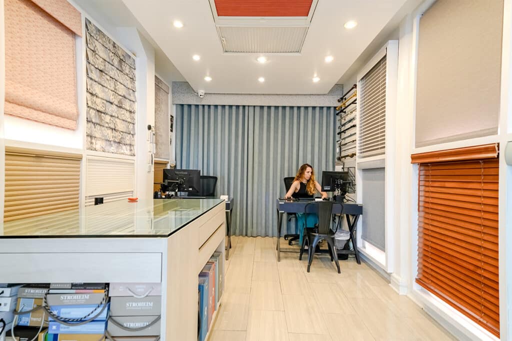 A woman sits at a desk in a modern showroom with various window blinds displayed on the walls. The room is brightly lit with recessed ceiling lights and has light-colored flooring and contemporary decor.