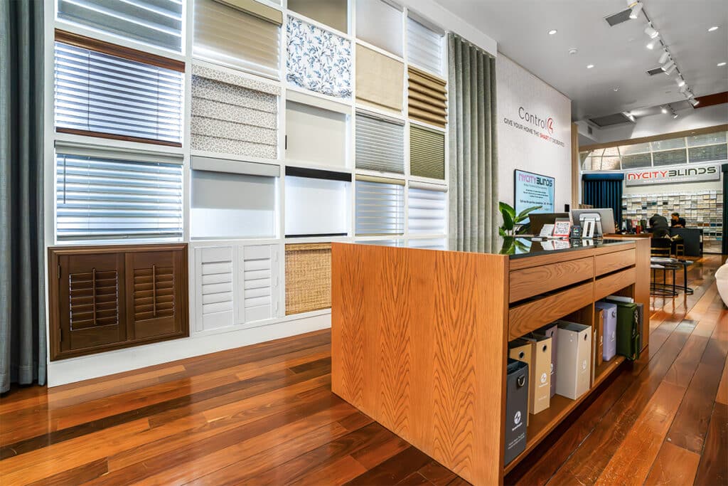 A modern showroom displays various window blinds and shades in a wall grid. A wooden desk with samples and plants stands on a polished hardwood floor. People talk at a seating area in the background.