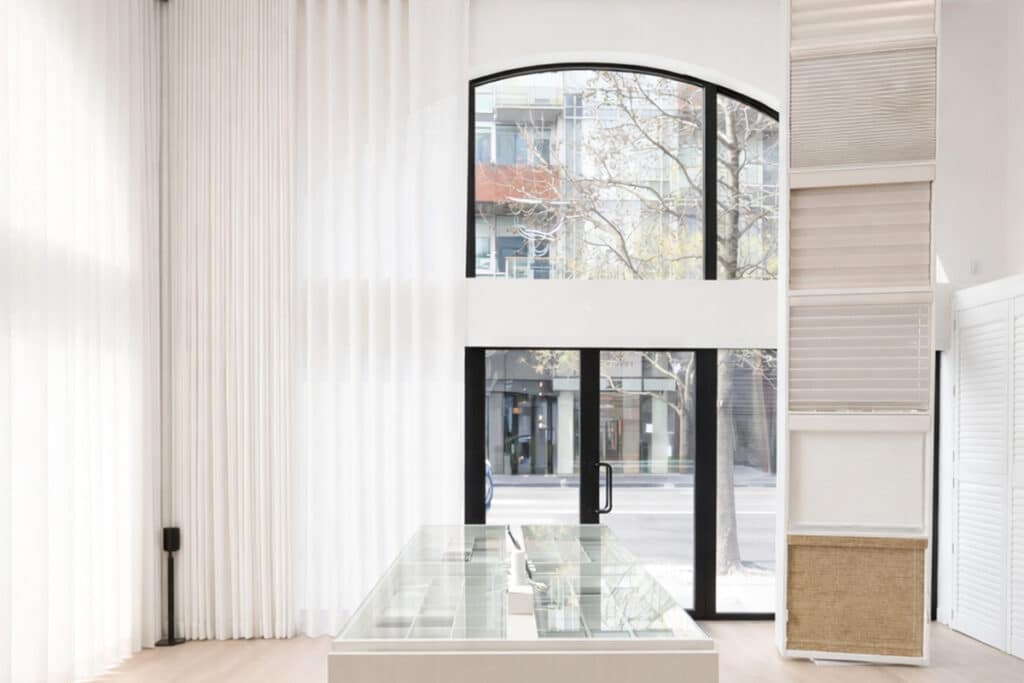 A bright, modern showroom with large arched windows, sheer white curtains, a glass display table, and a vertical rack displaying various window blind samples. City buildings are visible through the windows.
